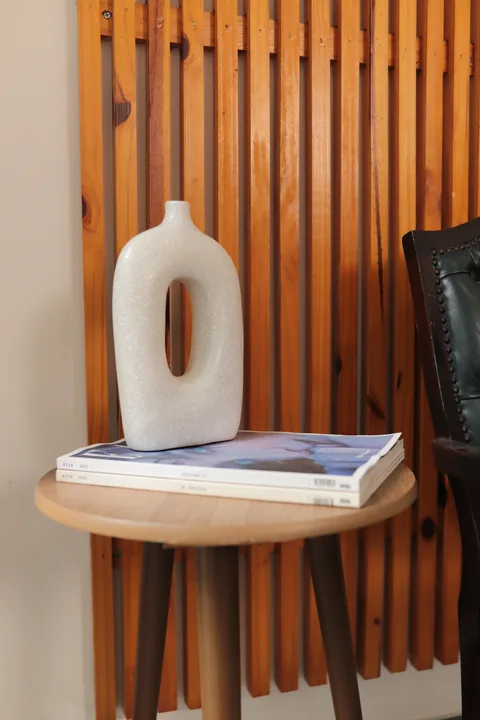 Vaso escultural branco sobre livros em mesa redonda, ao lado do painel ripado