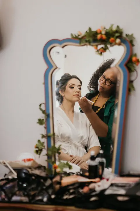Noiva sendo maquiada diante do espelho decorado com flores no camarim