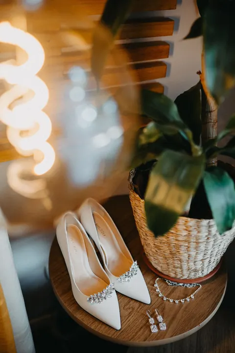 Sapatos de noiva com brilho e joias sobre mesa de madeira, ao lado de planta e luminária